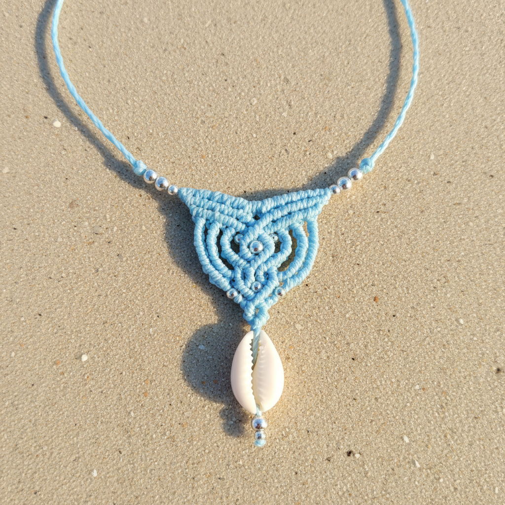Handmade micro macramé necklace with baby blue thread, cowrie shell drop and silver beads on a sandy coastal background